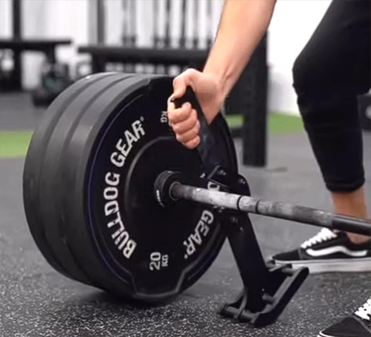 A close-up of a person's hand gripping a black handle on a barbell loaded with two black Bulldog Gear 20 kg weight plates, set on a gym floor.
