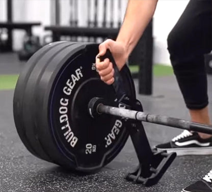 A close-up of a person's hand gripping a black handle on a barbell loaded with two black Bulldog Gear 20 kg weight plates, set on a gym floor.